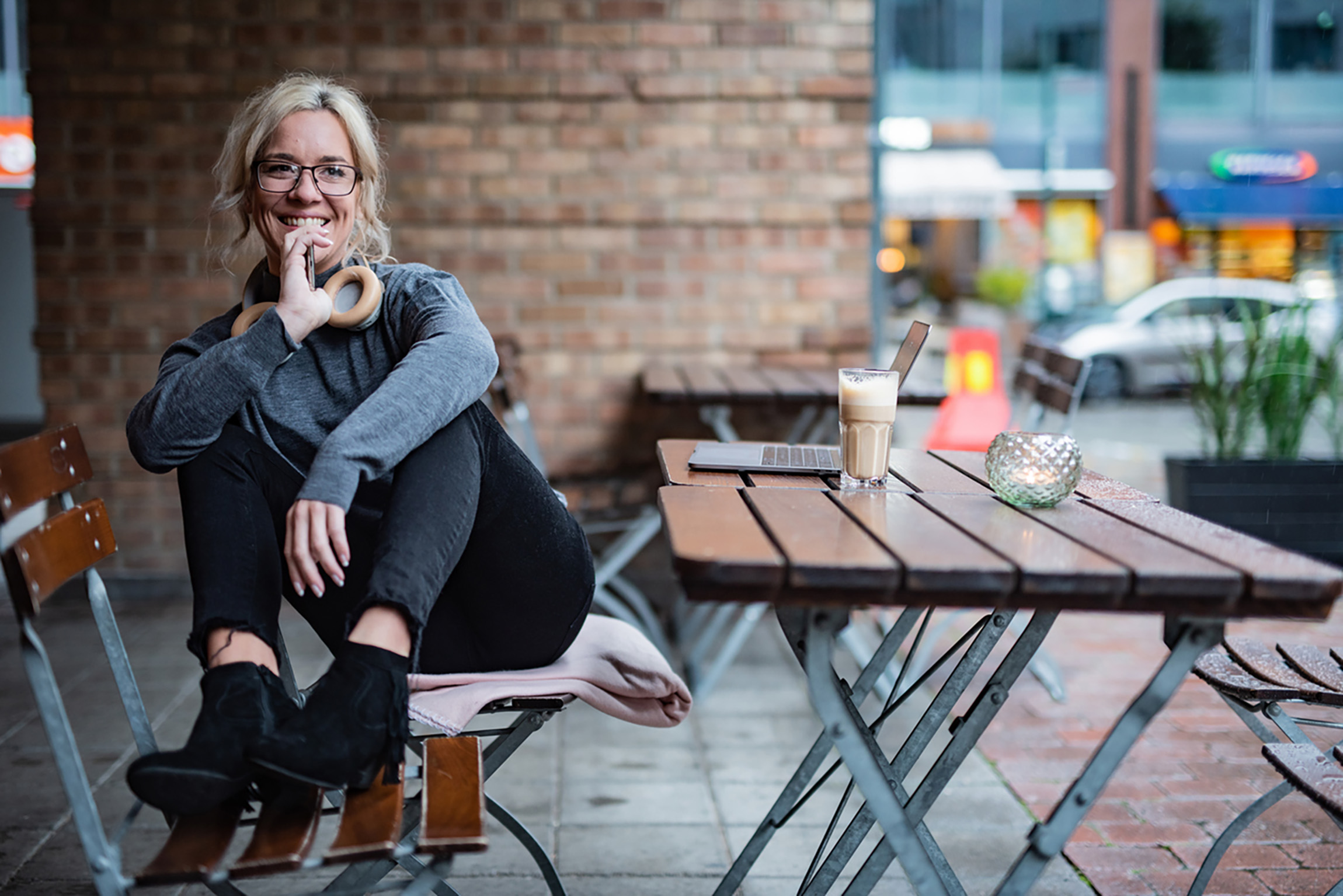 A young woman with headphones around her neck, sitting outside at a coffee bar