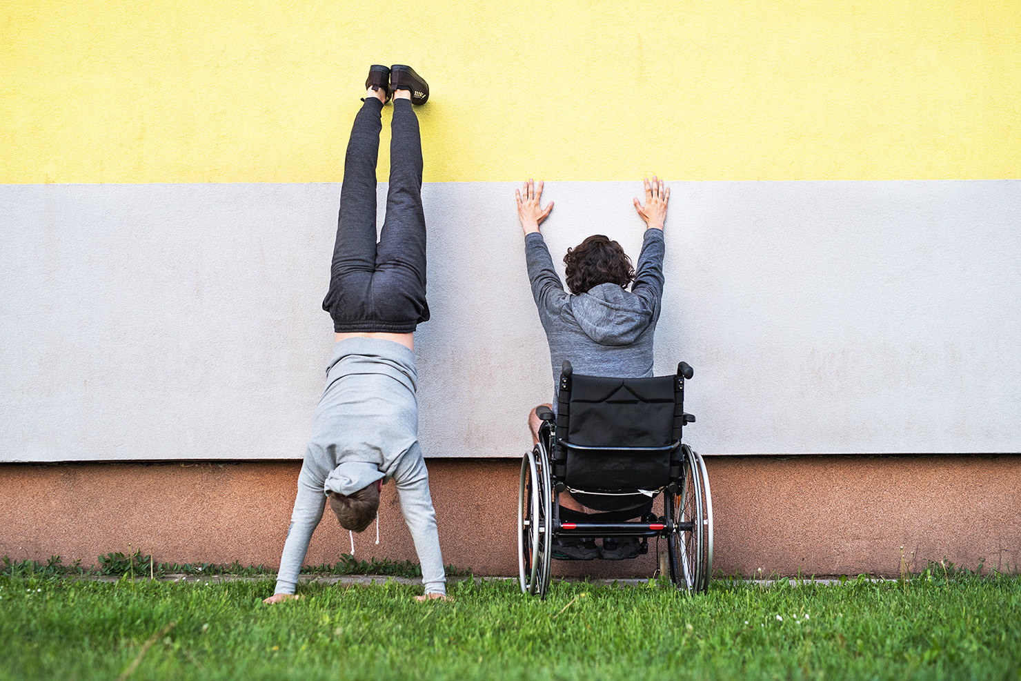 Two boys by a wall, one doing a handstand, one in a wheelchair with hands up